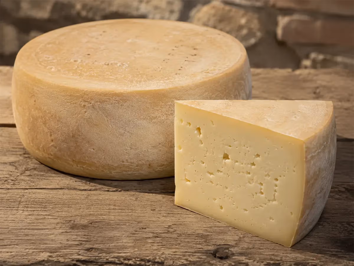 Forma intera di Asiago DOP con crosta beige chiara e spicchio separato su tavolo in legno grezzo