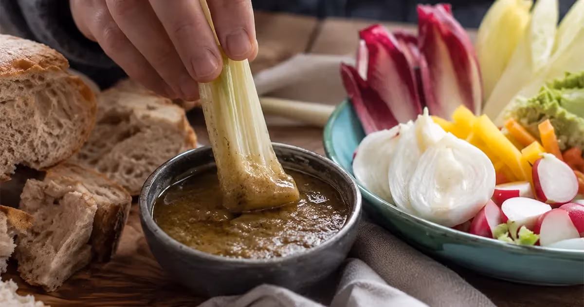 Mano che intinge un gambo di sedano nella bagna cauda calda color ocra servita in ciotola di terracotta scura affiancata da verdure fresche e pane rustico