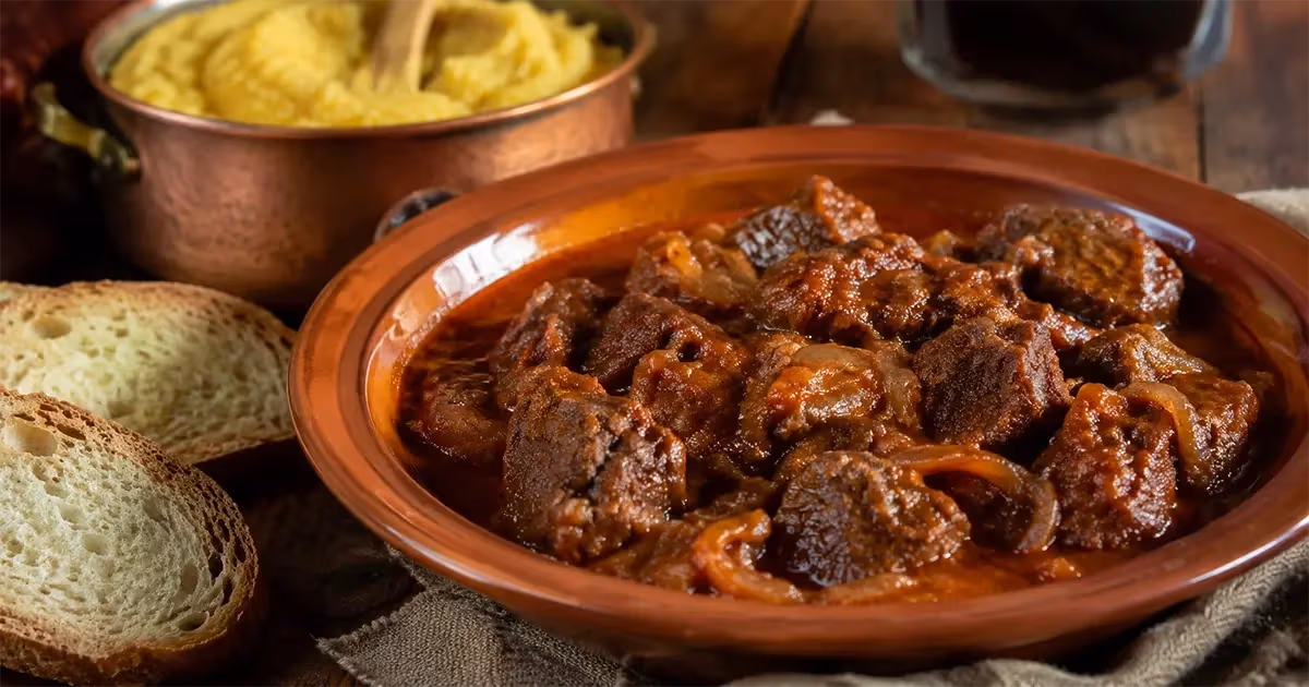 Goulash friulano in terrina di cotto con pezzi di carne brasata in sugo rosso scuro accompagnato da polenta gialla in pentola di rame e fette di pane rustico