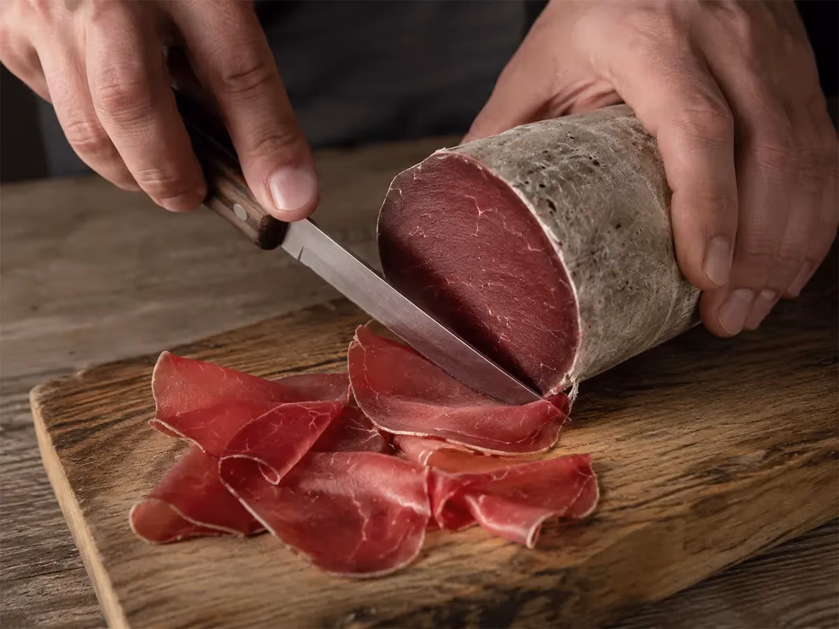 Mani che affettano bresaola della Valtellina con coltello su tagliere rustico in legno