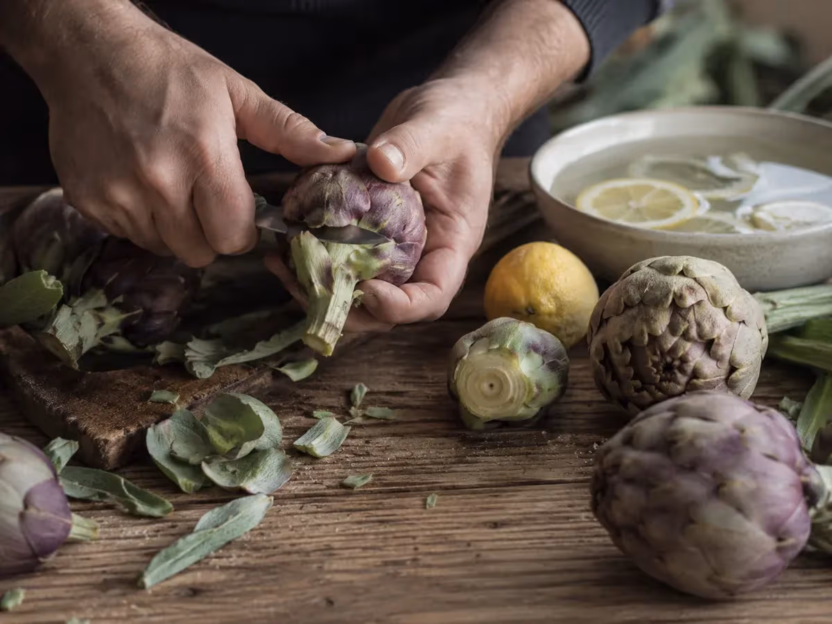 Mani che puliscono carciofi viola su tavolo di legno con limoni e ciotola di acqua acidulata