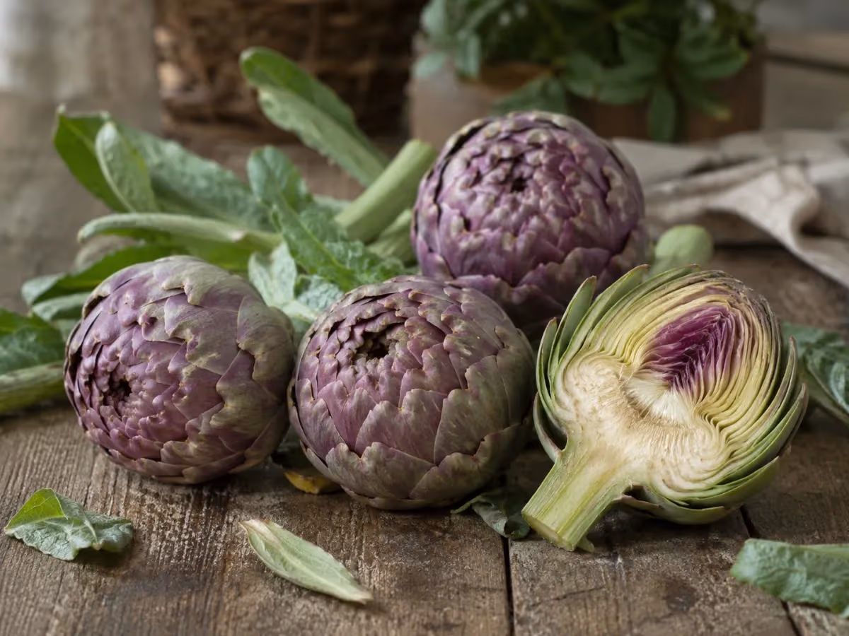 Quattro carciofi viola freschi su tavolo rustico uno tagliato a metà rivela l interno chiaro