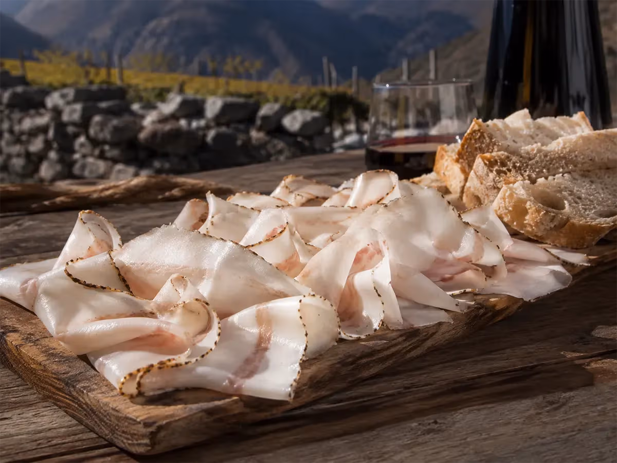 Fette trasparenti di lardo di Arnad DOP su tagliere rustico con pane e vino rosso in valle alpina