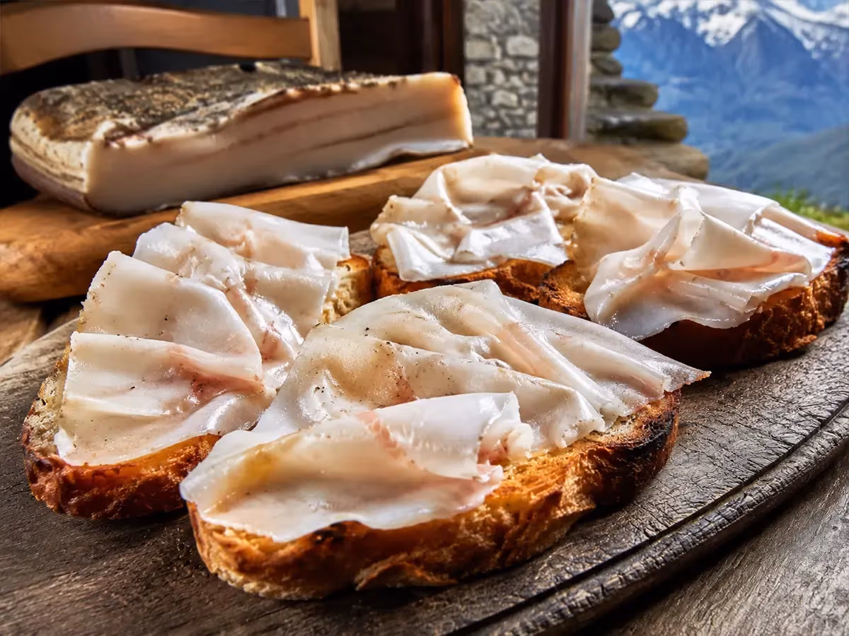 Bruschette di pane tostato ricoperte di lardo di Arnad DOP affettato con vista sulle Alpi innevate