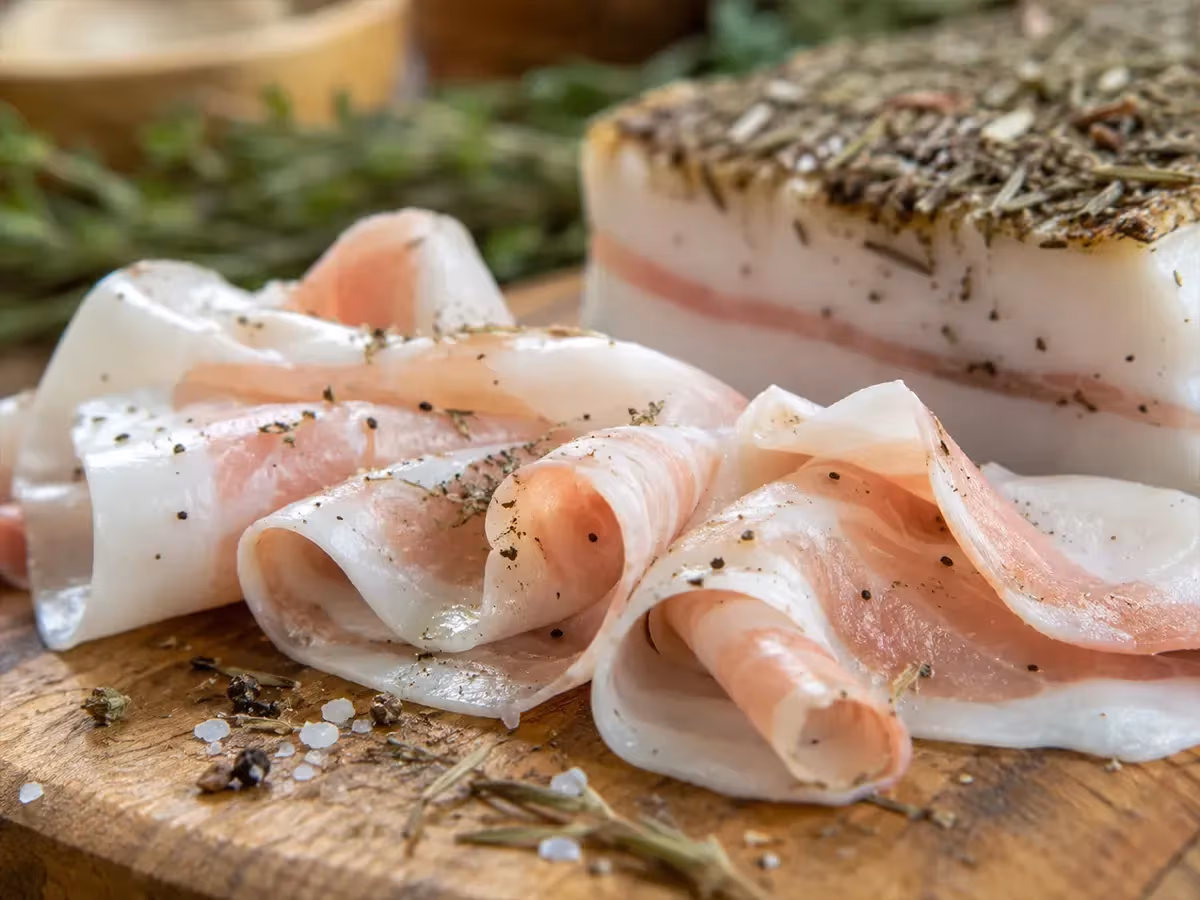 Fette arrotolate di lardo bianco con venature rosa su tagliere con rosmarino e grani di pepe