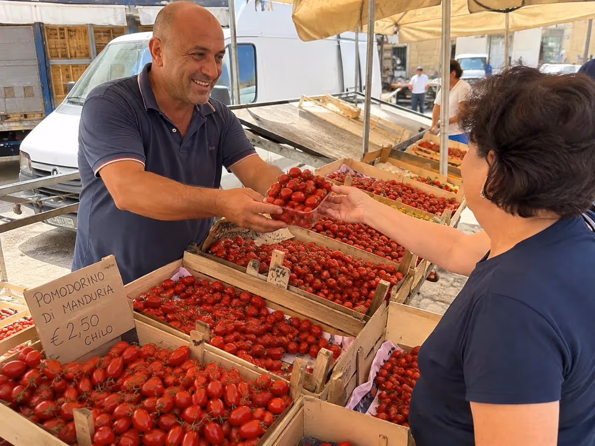 Venditore al mercato pugliese porge vaschetta di pomodorino di Manduria a cliente con cartello prezzo