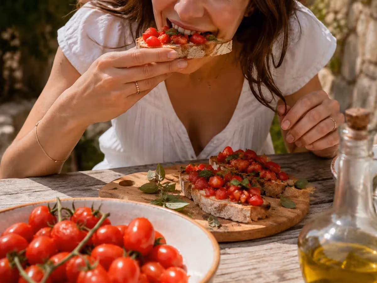 Donna morde una bruschetta con pomodori datterino tritati e basilico accanto a olio e piatto di pomodori freschi