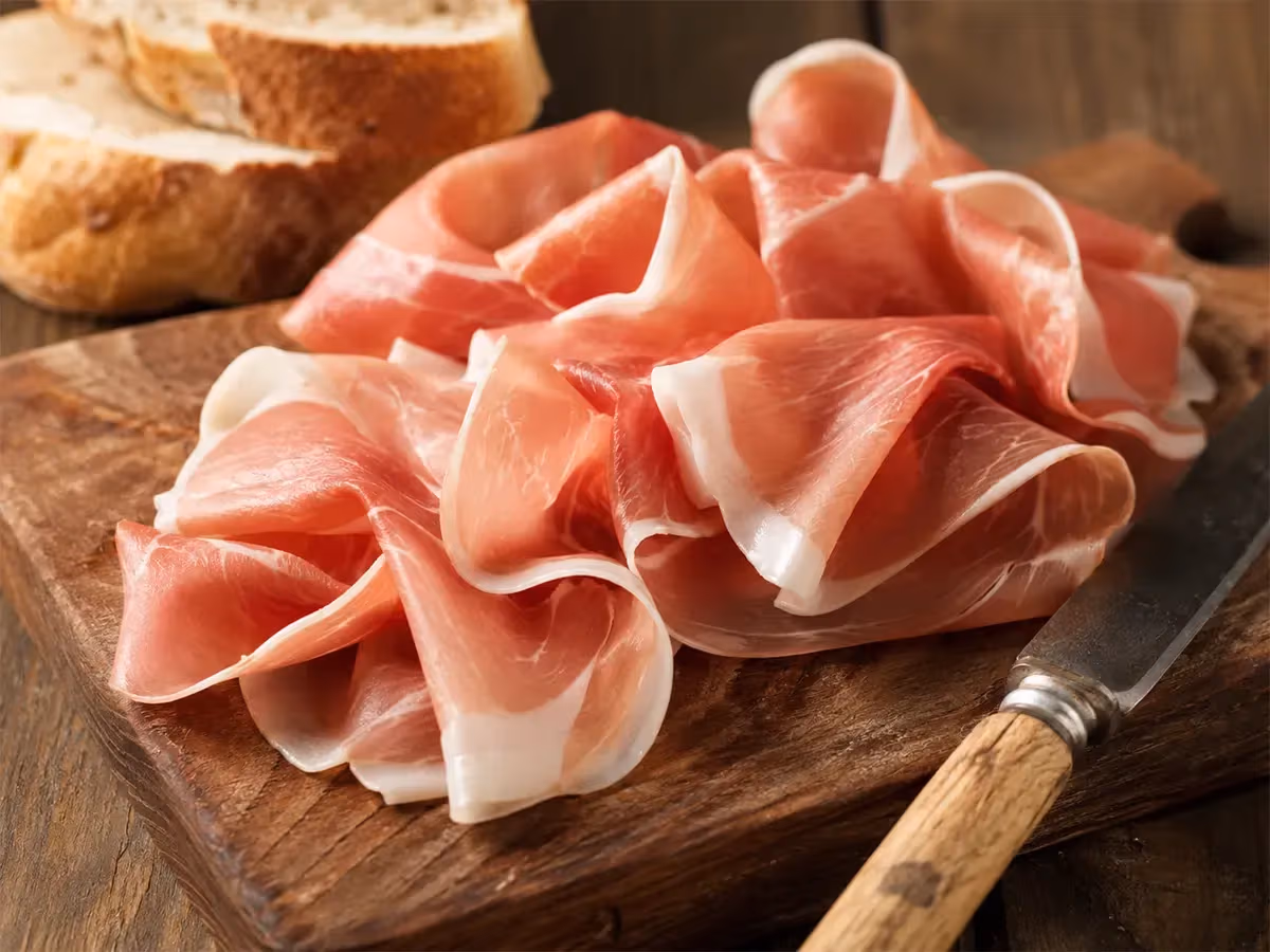 Fette sottili di prosciutto crudo rosa adagiate su tagliere di legno scuro con pane rustico