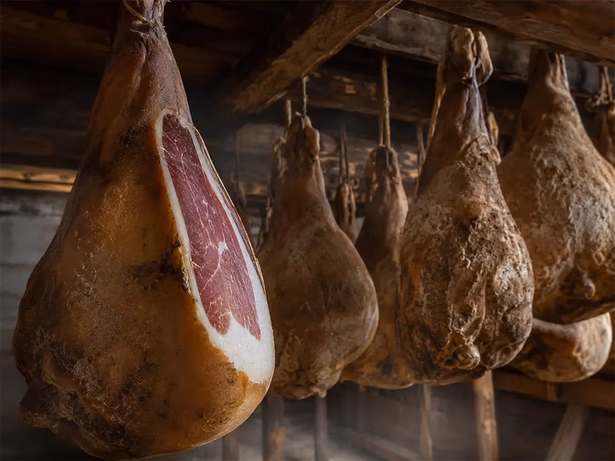 Prosciutti interi stagionati appesi in cantina buia con crosta scura e polpa rossa visibile